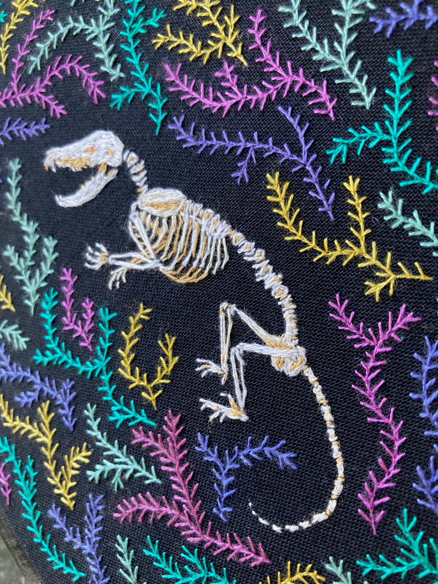 An embroidered opossum skeleton rests among colorful pink and teal and golden stitched moss or fern against a black background. 