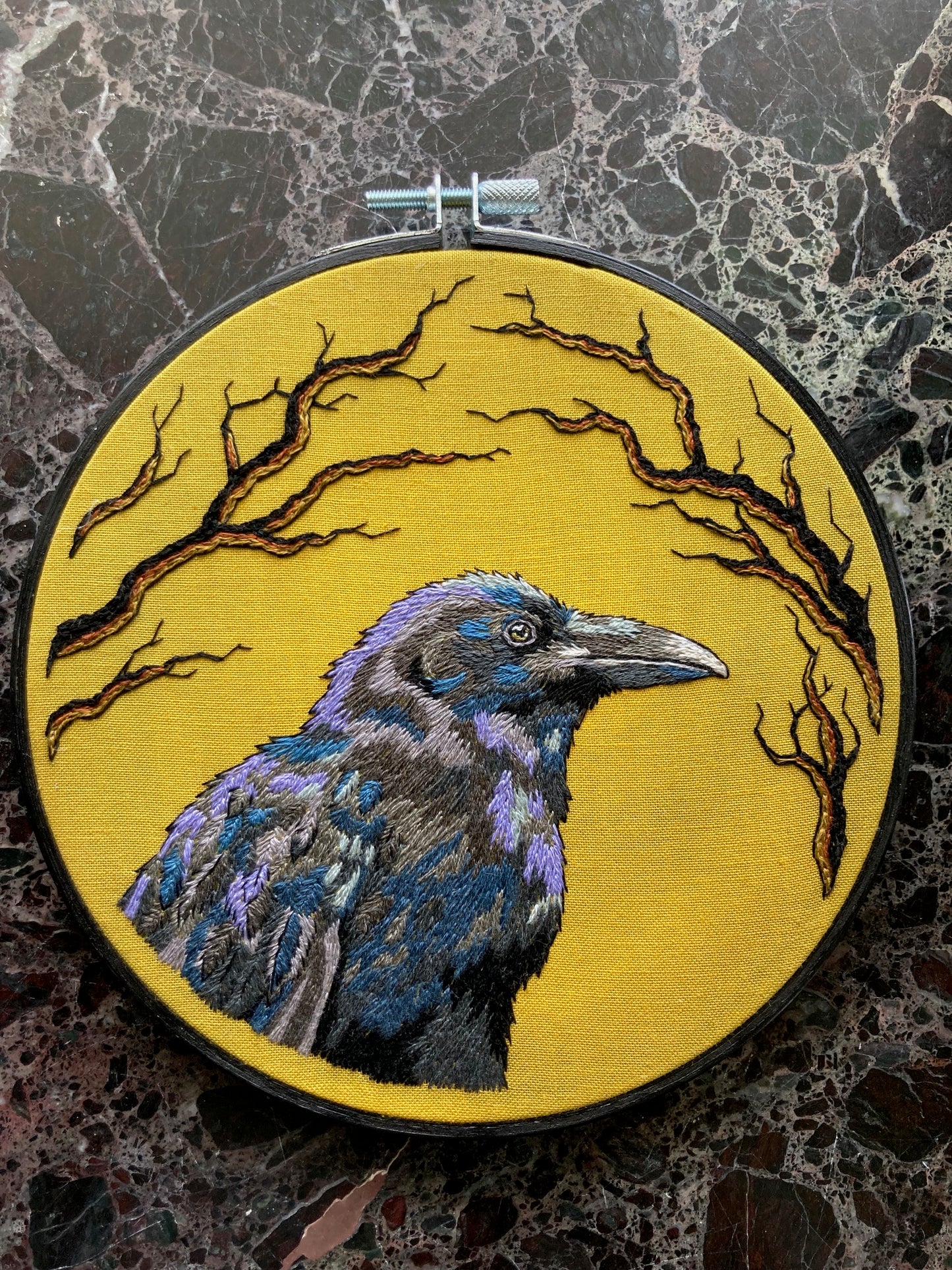 An embroidered raven stitched in black and purple threads faces to the right against a bright yellow background stitched with black branches around the edges, with a black painted round hoop