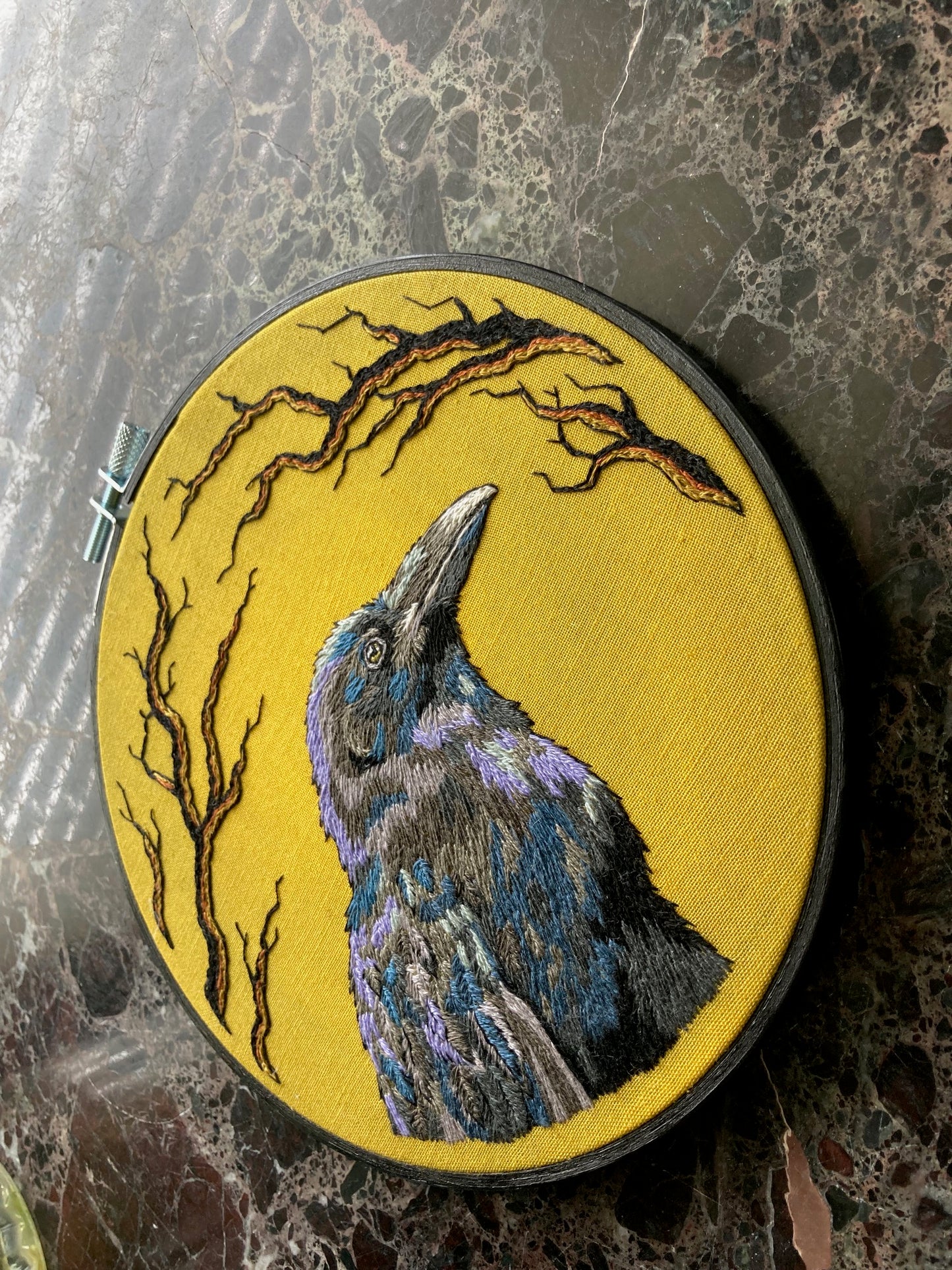 An embroidered raven stitched in black and purple threads faces to the right against a bright yellow background stitched with black branches around the edges, with a black painted round hoop