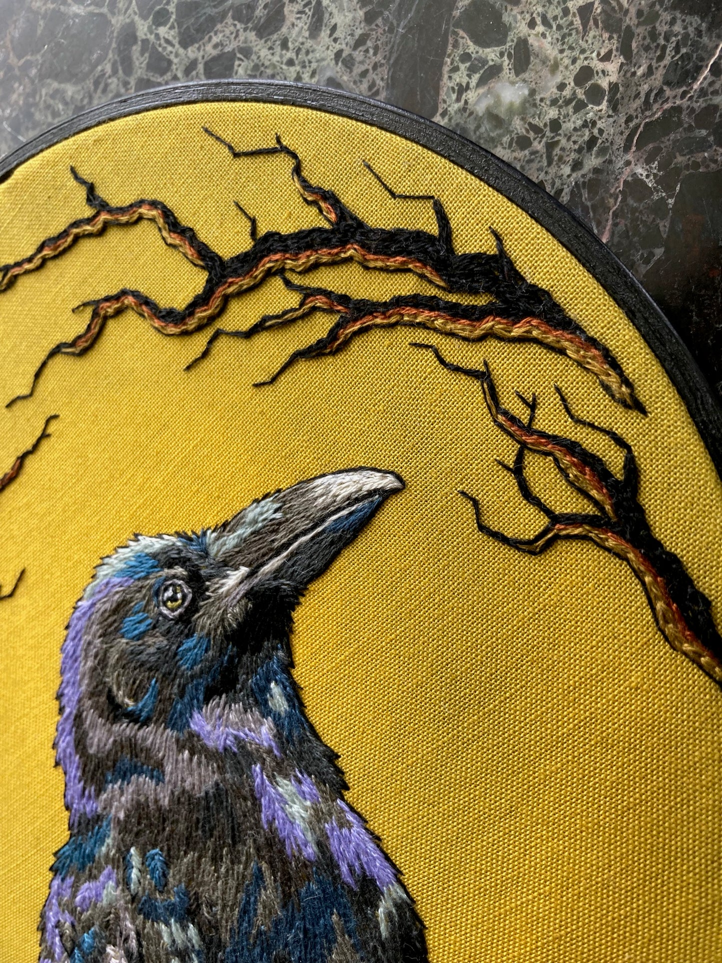 An embroidered raven stitched in black and purple threads faces to the right against a bright yellow background stitched with black branches around the edges, with a black painted round hoop