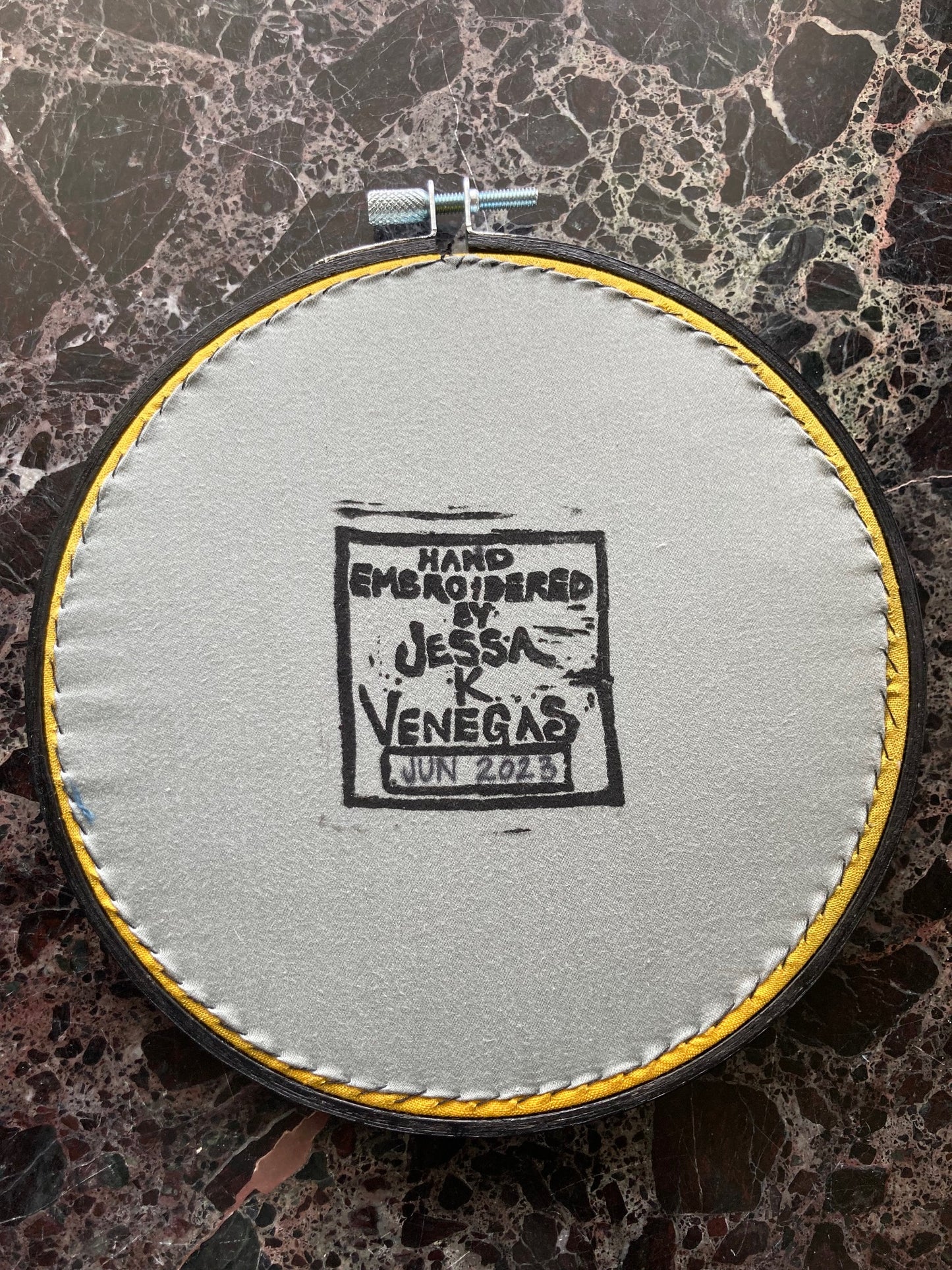 grey fabric is stitched tight into a black round embroidery hoop and printed with the words "Hand Embroidered by Jessa K Venegas" on it. June 2023 is written in ink. 