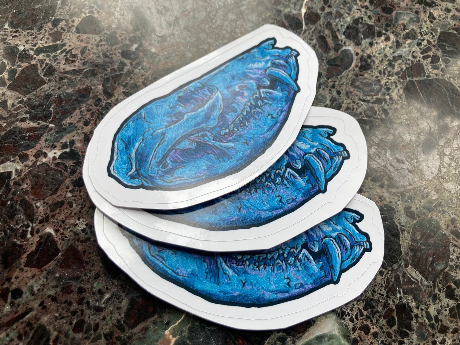 a pile of stickers with an image of an embroidered opossum skull in blue and purple tones sits on a dark surface. 