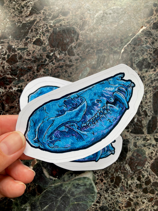 a pile of stickers with an image of an embroidered opossum skull in blue and purple tones sits on a dark surface. A hand is holding one up to the camera.