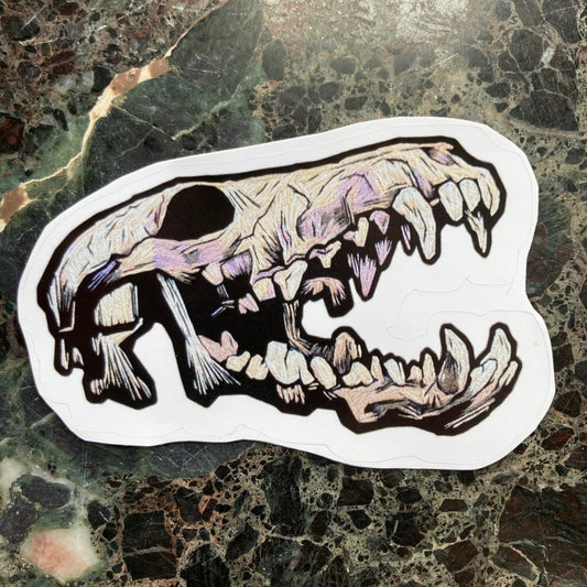 a sticker depicting an embroidered coyote skeleton with an open mouth and lots of teeth sits on a dark surface. The Embroidered skull is beige and lavender and black tones with lots of shadows.
