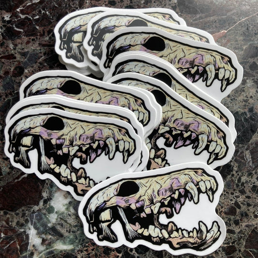 a pile of stickers depicting an embroidered coyote skeleton with an open mouth and lots of teeth sits on a dark surface. The Embroidered skull is beige and lavender and black tones with lots of shadows.