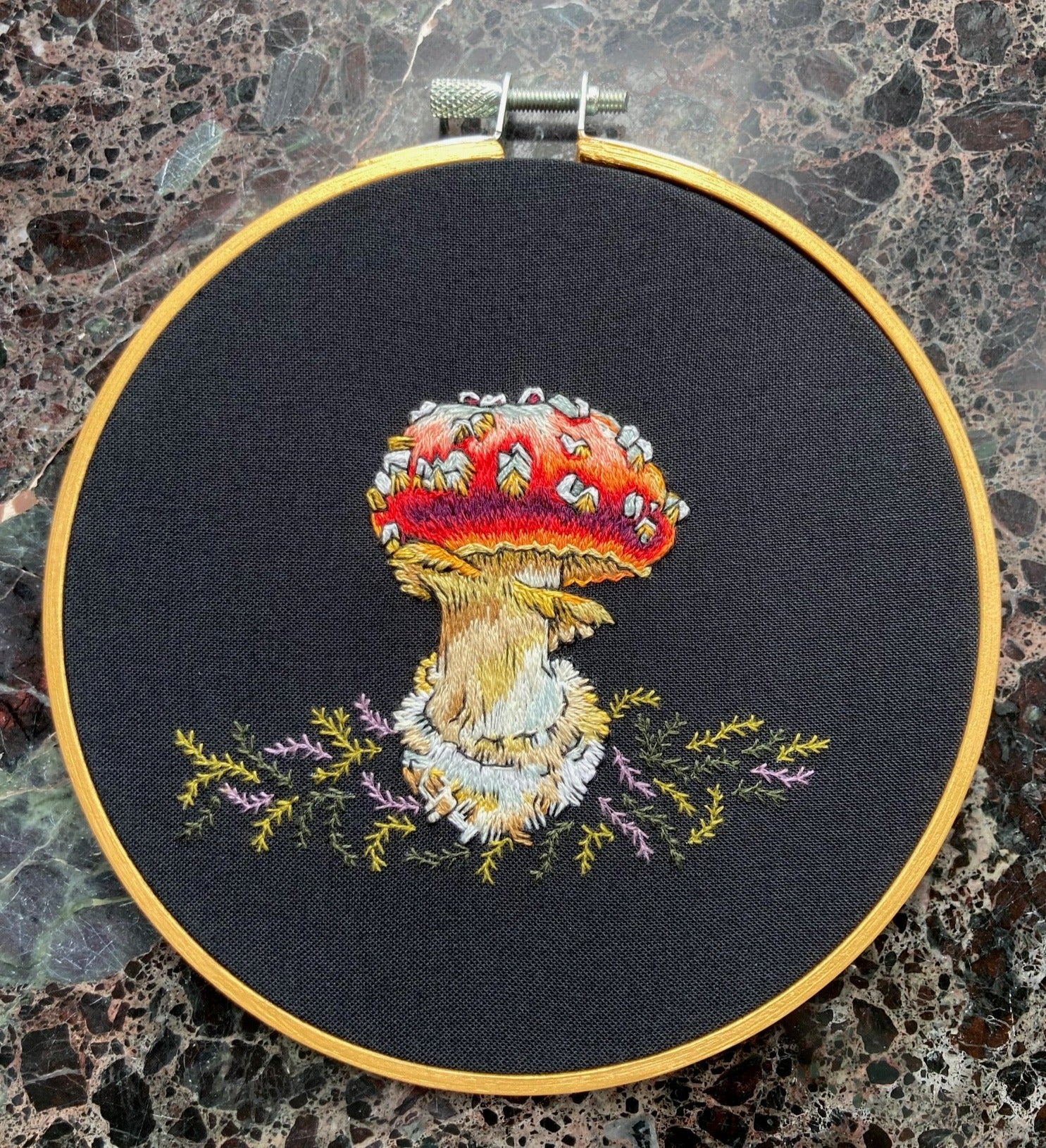 A brightly colored amanita mushroom with a red cap is embroidered on black fabric with green and lavender moss. The fabric is stretched into a gold round embroidery hoop. 