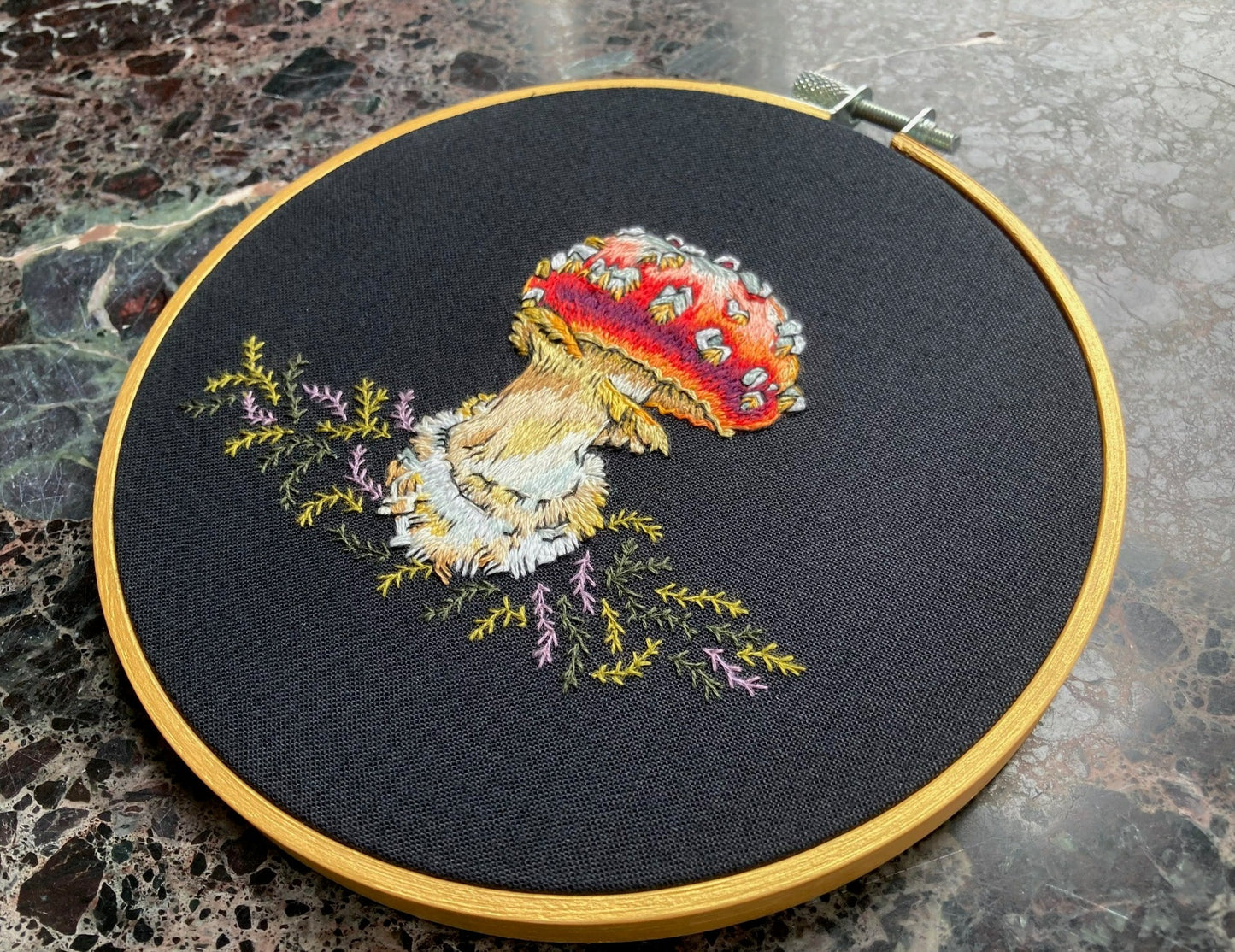 A brightly colored amanita mushroom with a red cap is embroidered on black fabric with green and lavender moss. The fabric is stretched into a gold round embroidery hoop. The picture is taken at an extreme angle to catch the light on the threads. 