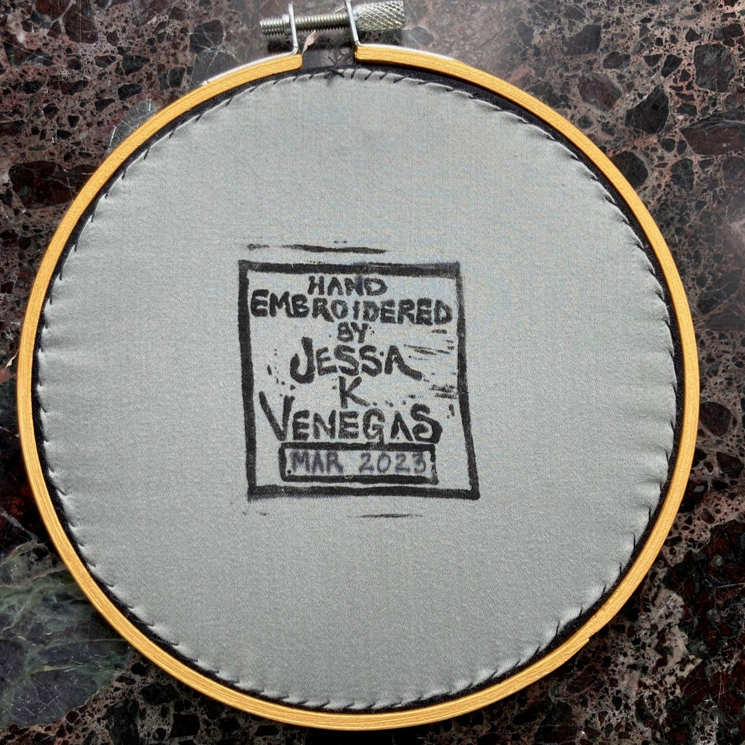 Grey fabric is stitched taut onto the back of a gold round embroidery hoop. The fabric is printed with a label that says "Hand Embroidered by Jessa K Venegas" with the date March 2023 written in ink below it.