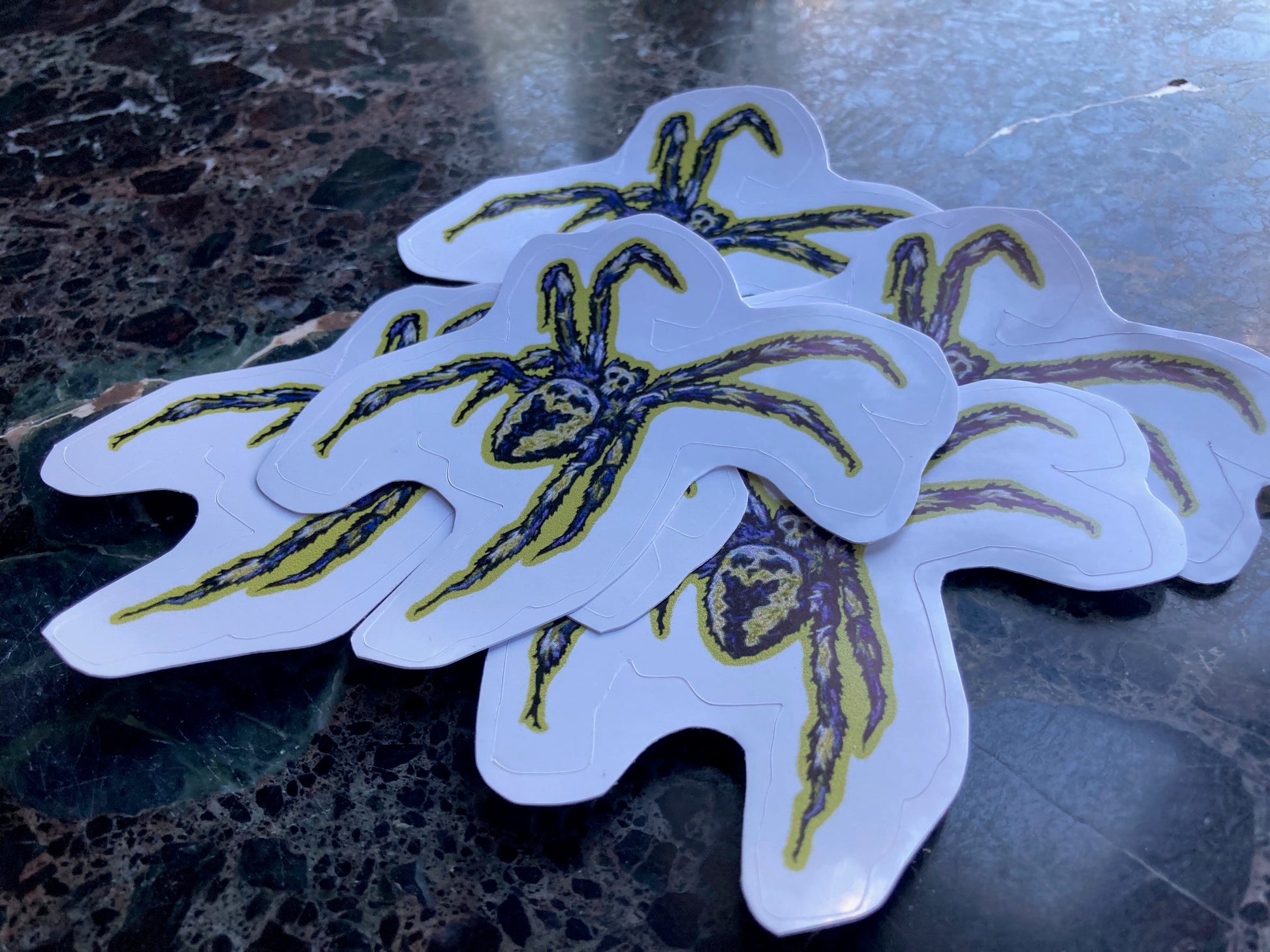 A pile of stickers sits on a dark surface. The stickers depict an embroidered fuzzy spider in deep purple and chartreuse and black, with the shape of  skull in its markings. 