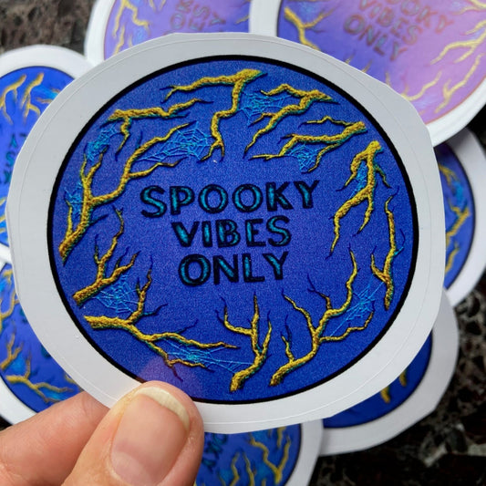 a pile of stickers lays on a dark surface. a hand is holding one up to the camera, depicting an embroidery of golden branches on a deep periwinkle blue background and the words SPOOKY VIBES ONLY in the center. 