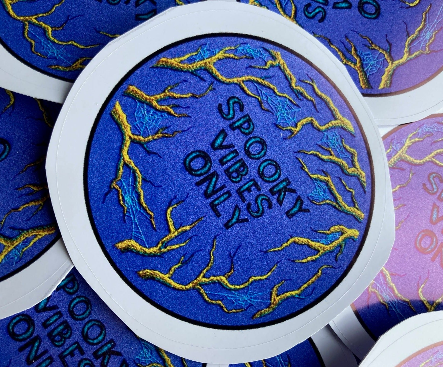 a pile of stickers lays on a dark surface. The stickers depict an embroidery of golden branches on a deep periwinkle blue background and the words SPOOKY VIBES ONLY in the center.
