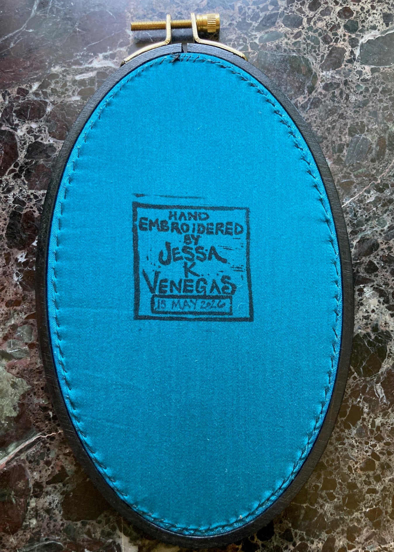the back of an embroidery hoop, with teal fabric stitched taut across it. The fabric is printed with HAND EMBROIDERED BY JESSA K VENEGAS and the date 15 May 2026