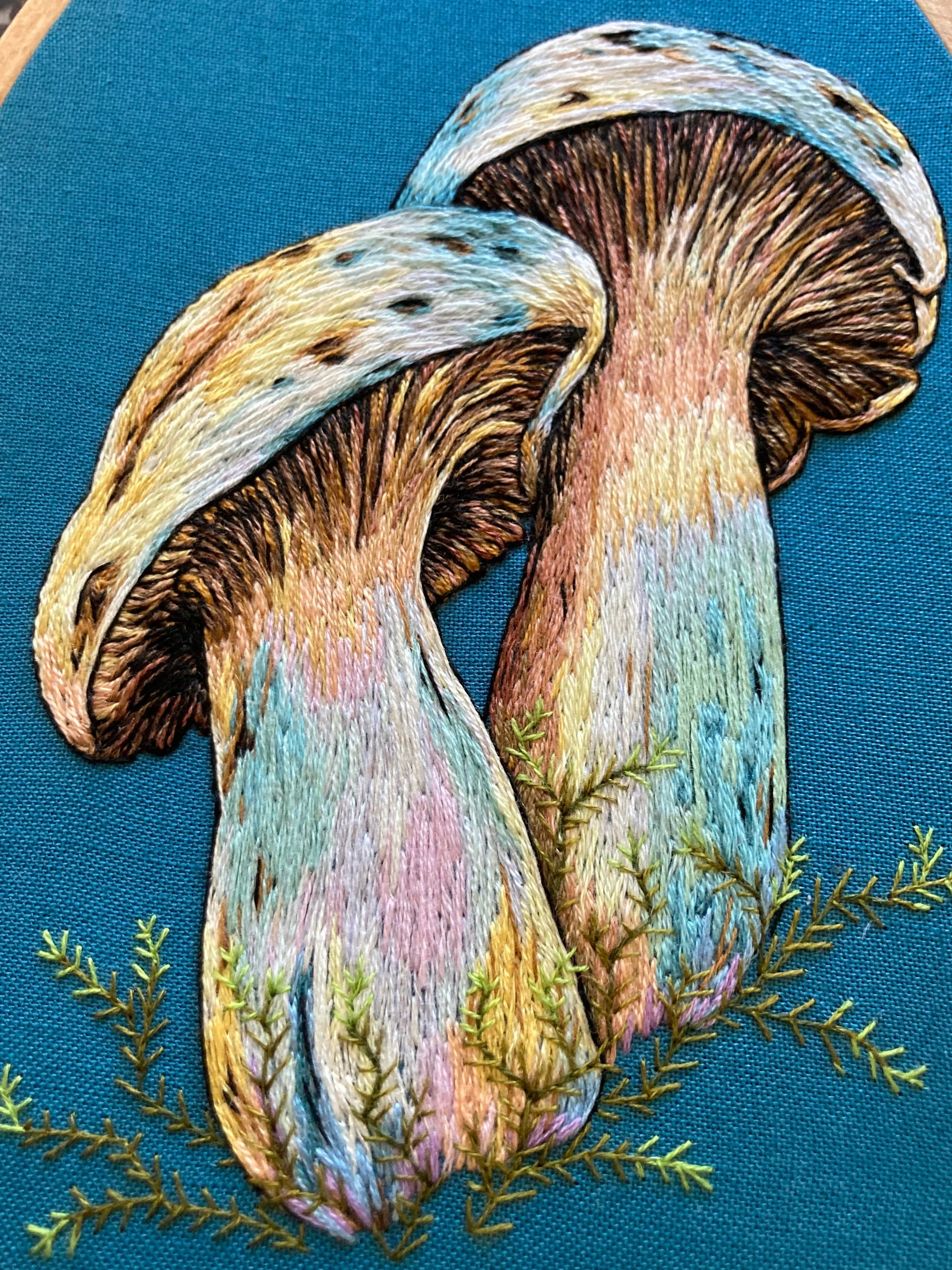 close up picture of a pair of colorful hand-embroidered mushrooms on teal fabric