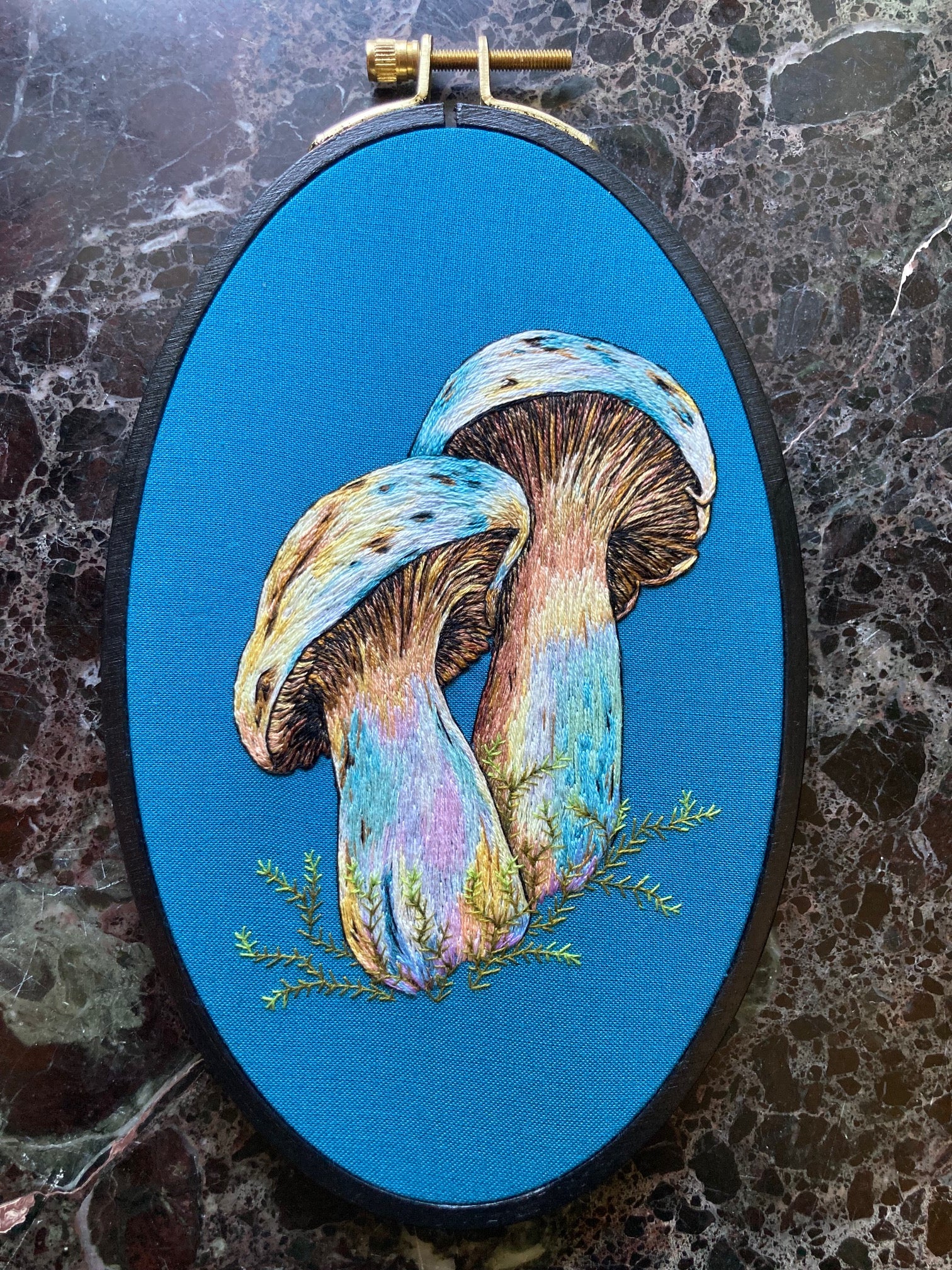 a pair of colorful hand-embroidered mushrooms on teal fabric, in a black embroidery hoop sitting on a black marble surface