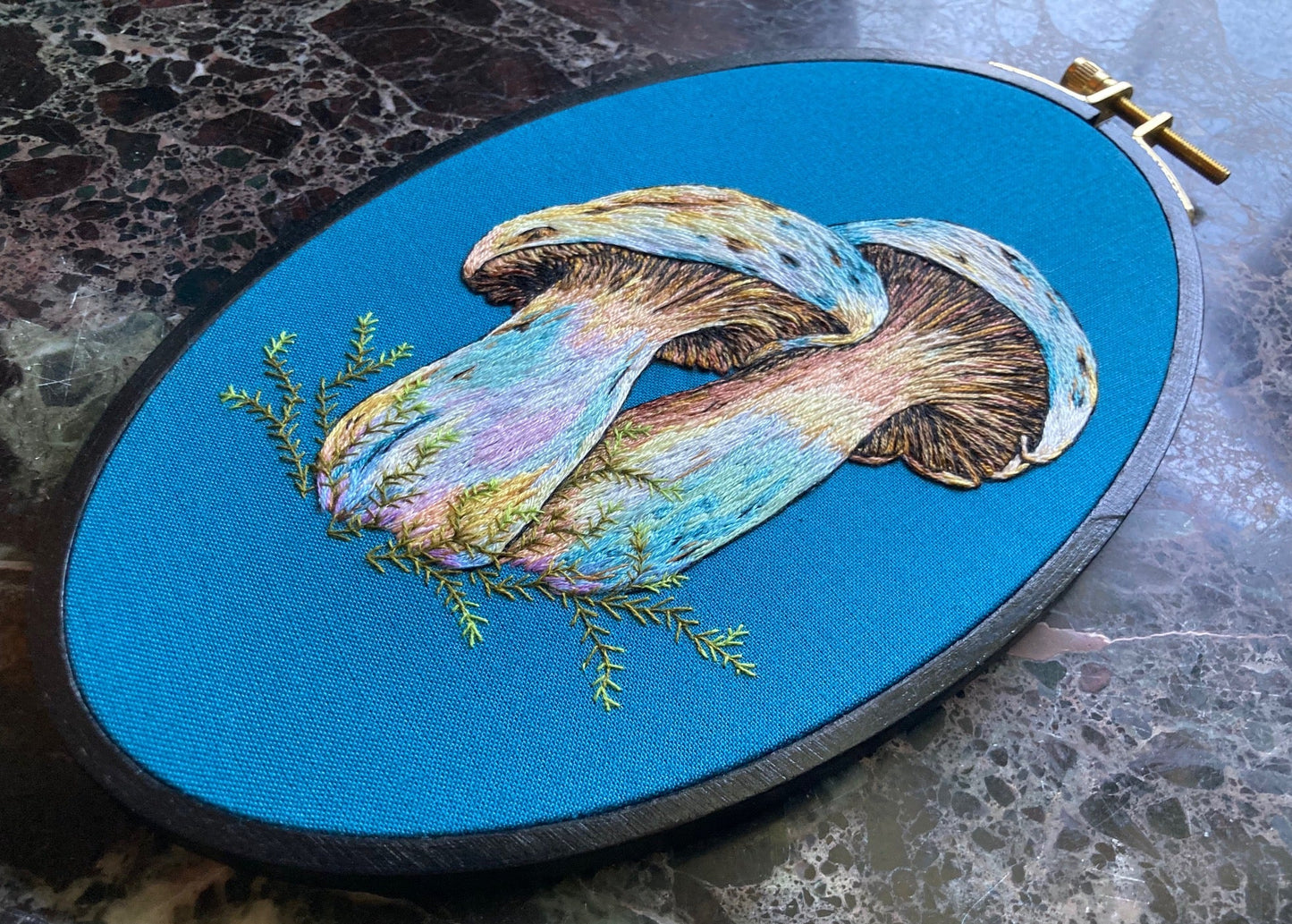 colorful hand-embroidered mushrooms on teal fabric, in a black embroidery hoop sitting on a black marble surface