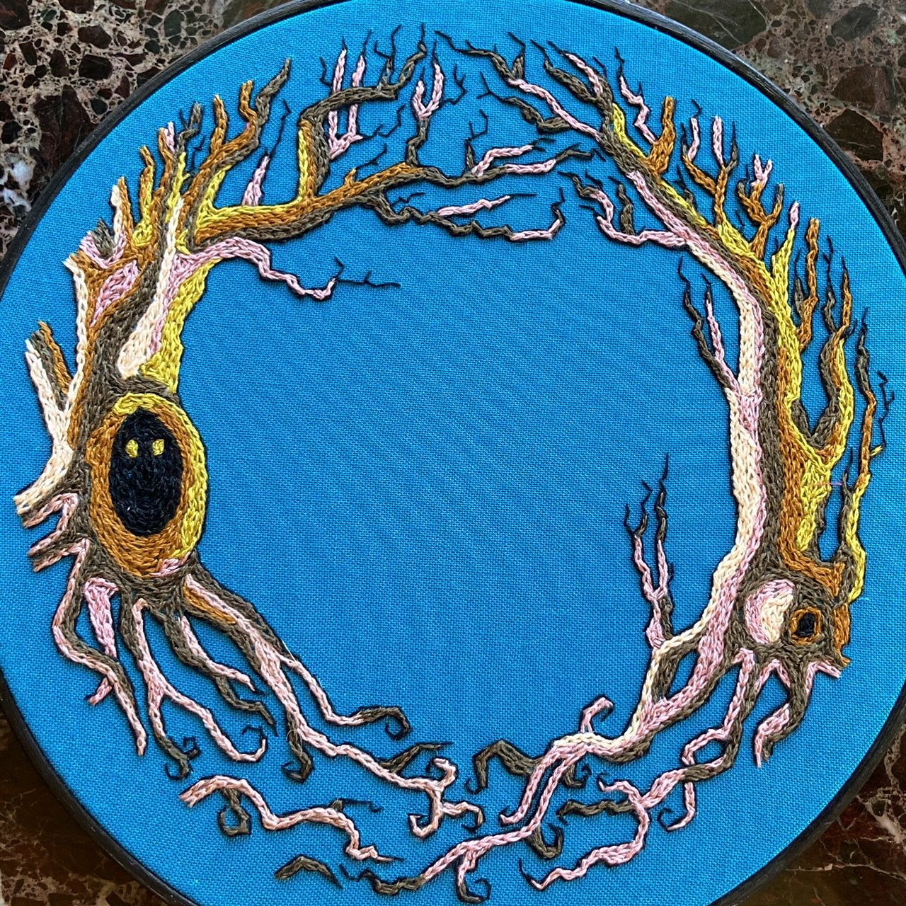 An embroidery hoop sits on a black marble background. It features two gnarled trees stitched in brown and lilac and green. One tree has little eyes peering out from a dark knot. 