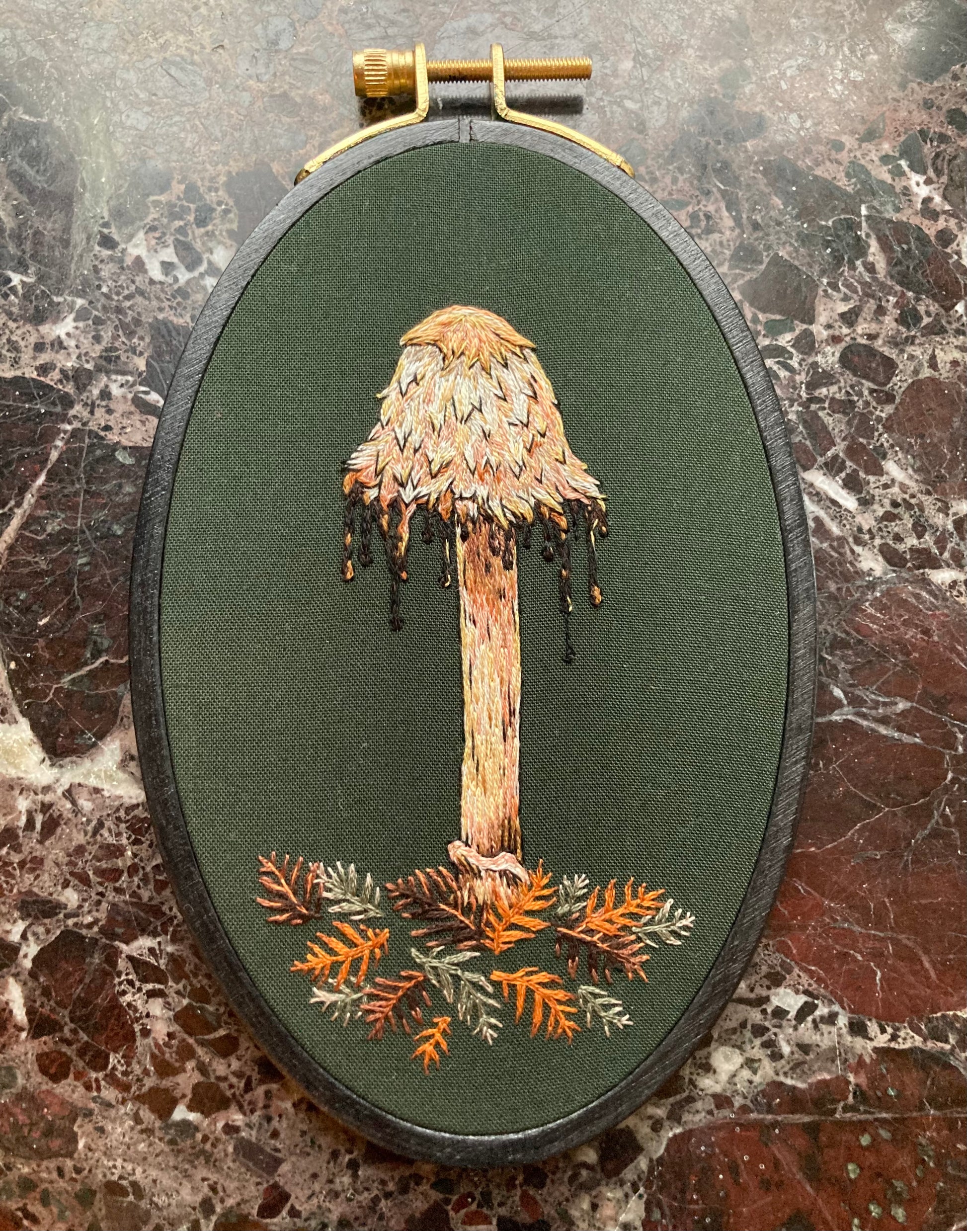 An embroidery hoop sits on a black marble background. It features a hand embroidered shaggy mane mushroom with a drippy cap and some stitched redwood leaves, on a dark green fabric.