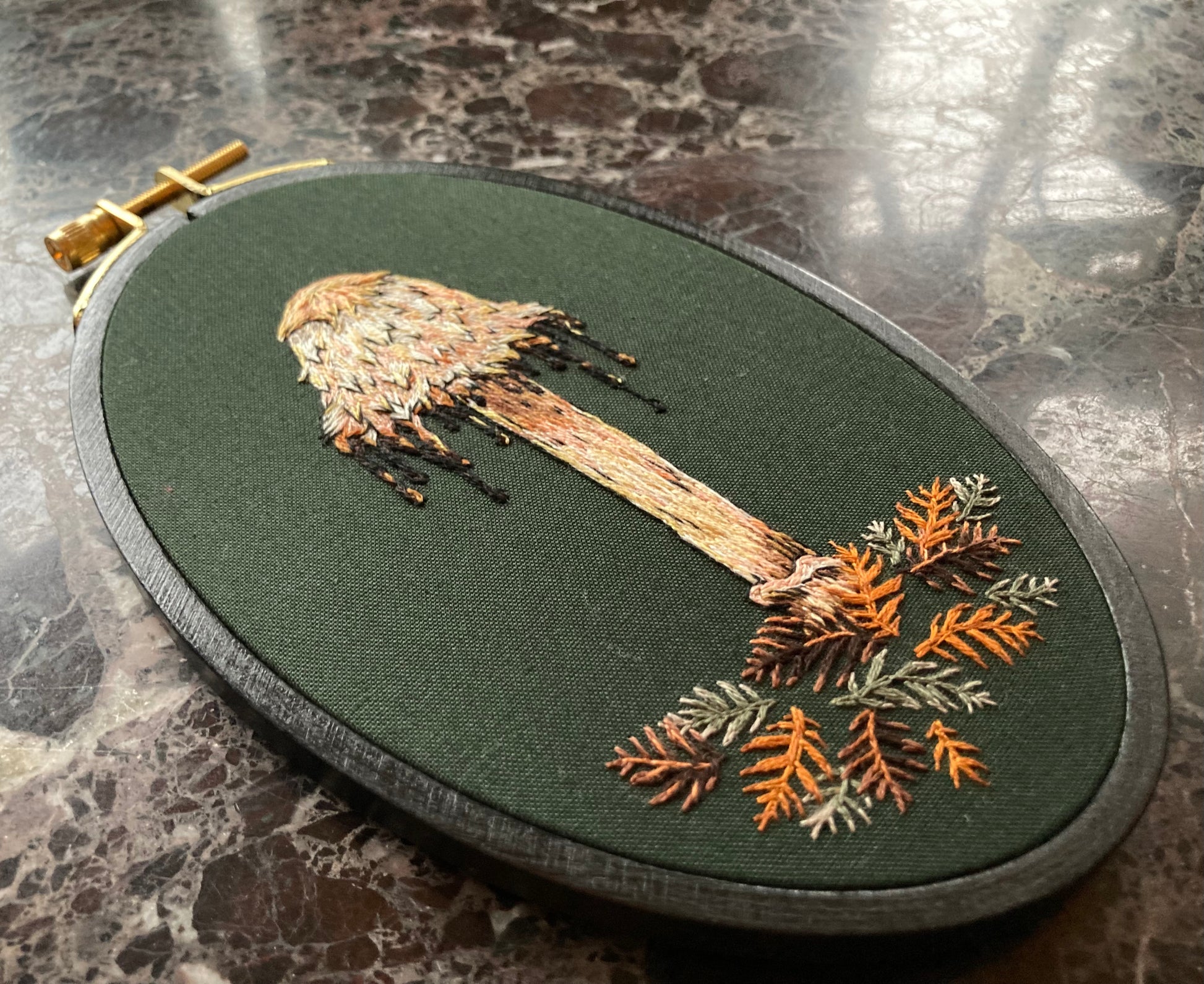 An embroidery hoop sits on a black marble background. It features a hand embroidered shaggy mane mushroom with a drippy cap and some stitched redwood leaves, on a dark green fabric. 