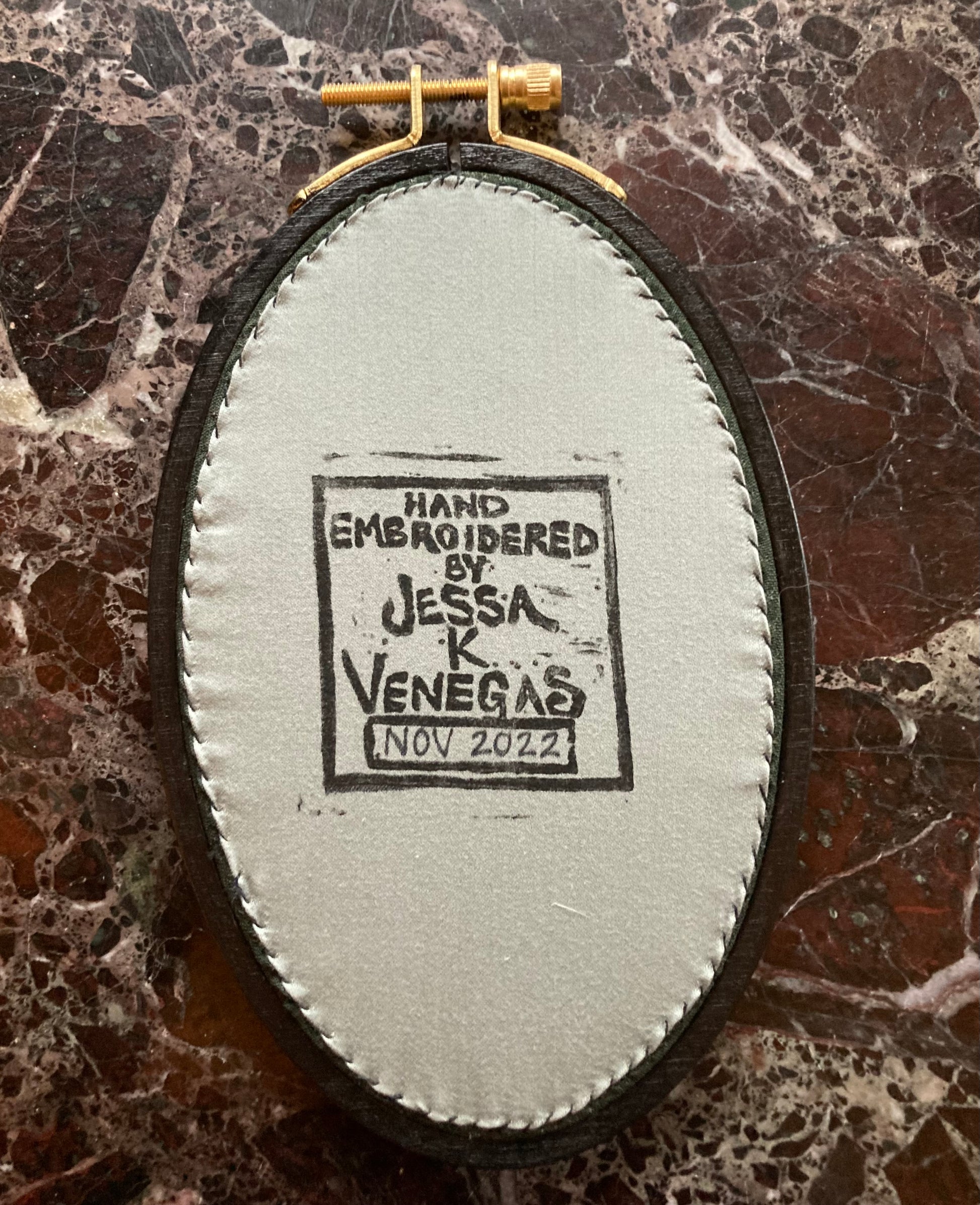 The back of an embroidery hoop with light grey fabric stitched taut like a drum. A stamp reads "HAND EMBROIDERED BY JESSA K VENEGAS NOV 2022"