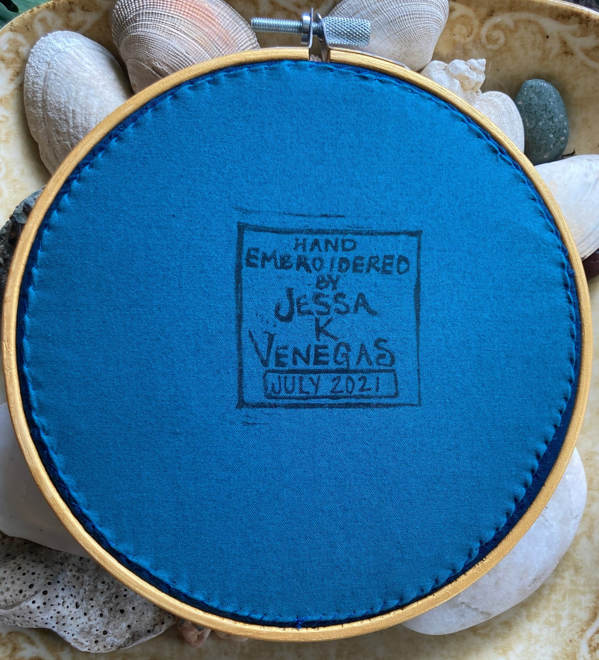 The back of an embroidery hoop with blue fabric stitched taut like a drum. A stamp reads "HAND EMBROIDERED BY JESSA K VENEGAS JUL 2021"