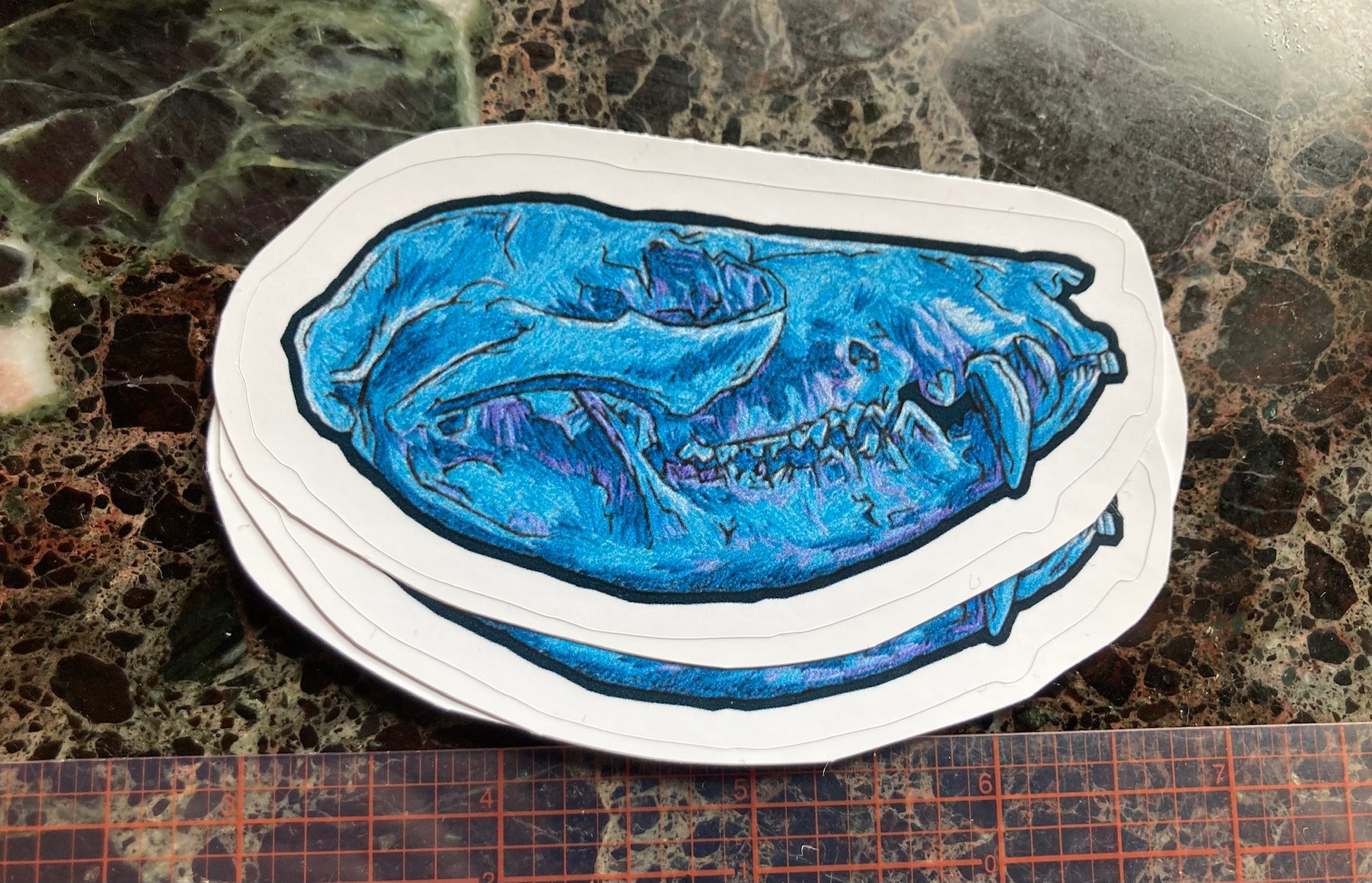 a pile of stickers with an image of an embroidered opossum skull in blue and purple tones sits on a dark surface. There is a visible ruler showing them to be a little less than 4 inches in length. 