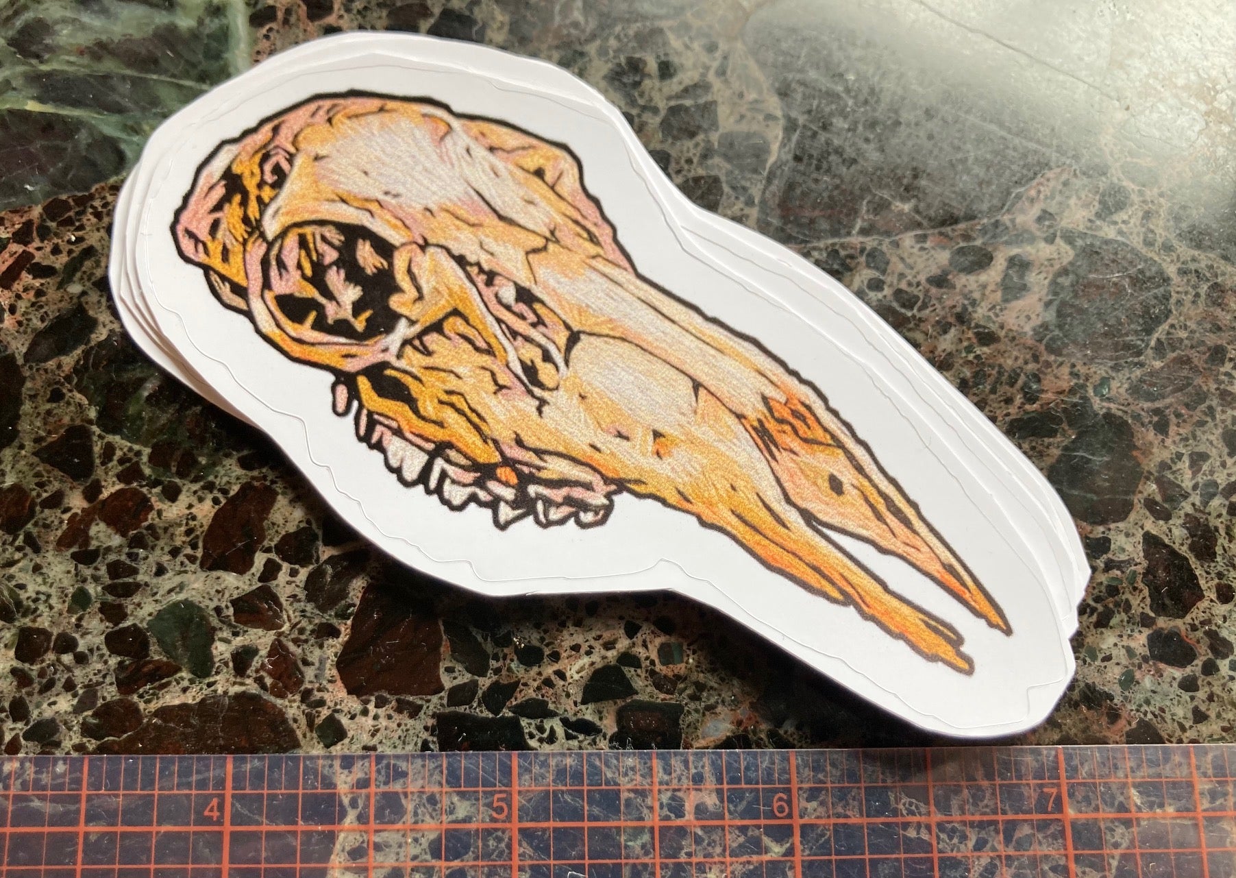 a sticker of an embroidered deer skull with no lower jaw sits on a dark surface. there is a visible ruler showing it is approximately 4 inches long. 