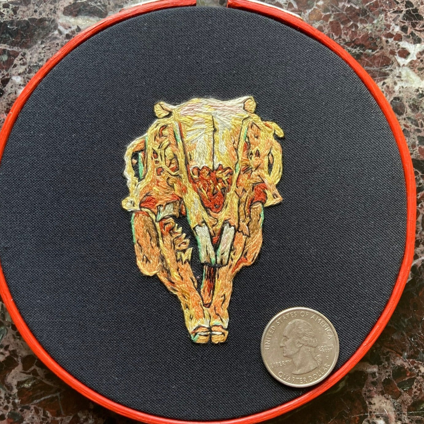 Hand embroidery of a rabbit skull on black fabric in a red, round hoop.  There is a quarter in the picture to show it compared to the 5" hoop size.