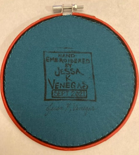 The back of an embroidery hoop with teal fabric stitched onto it tightly. The fabric has a printed label that says "HAND EMBROIDERED BY JESSA K VENEGAS SEPT 2021" and the artist signature.