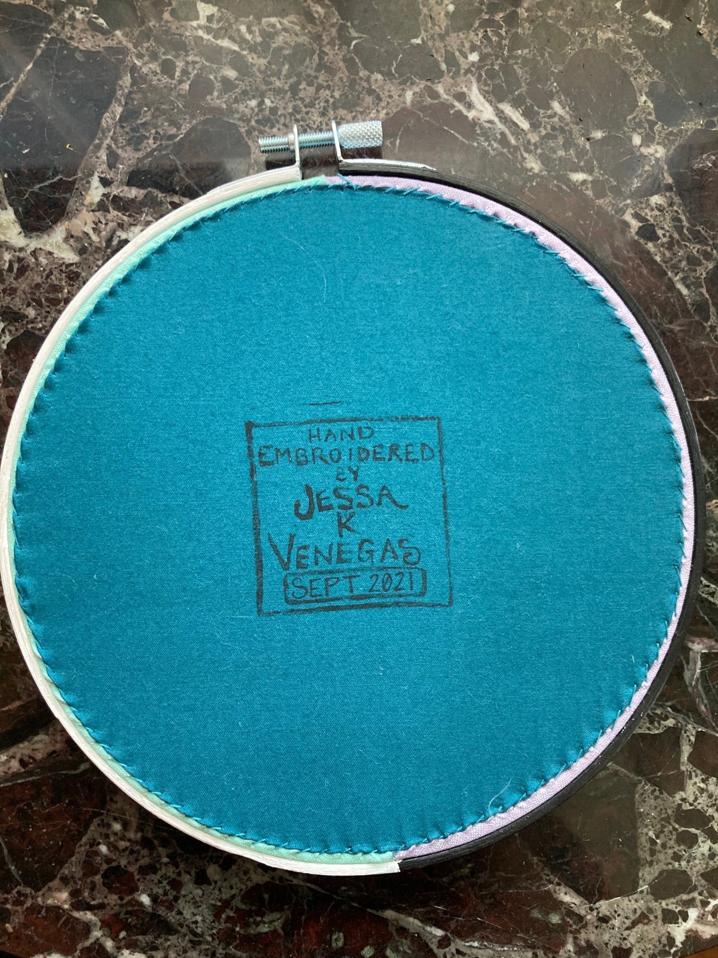 The back of an embroidery hoop with blue fabric stitched taut like a drum. A stamp reads "HAND EMBROIDERED BY JESSA K VENEGAS SEPT 2021"