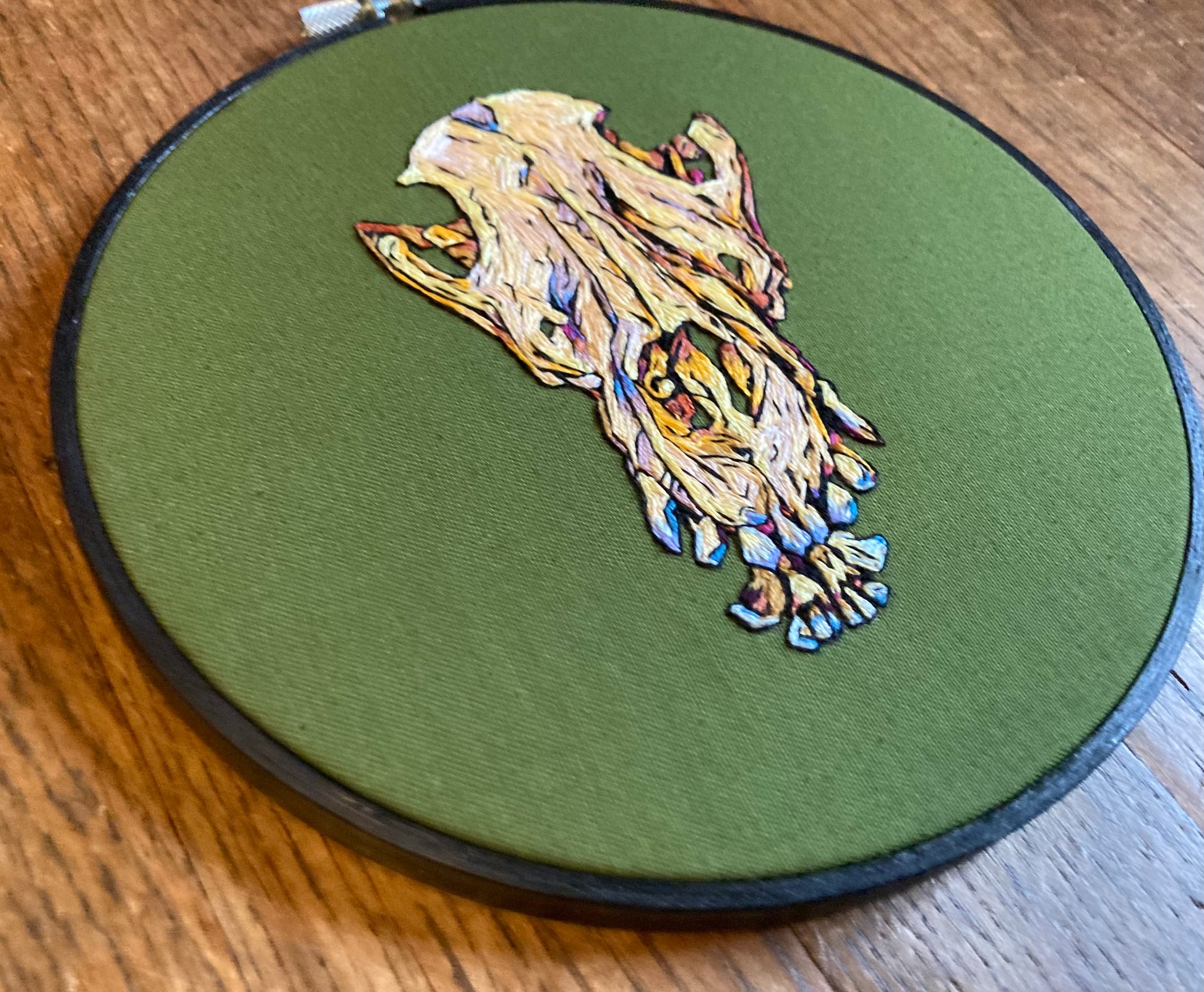 Colorful embroidery of a dog skull with prominent teeth on olive green fabric in a round embroidery hoop.