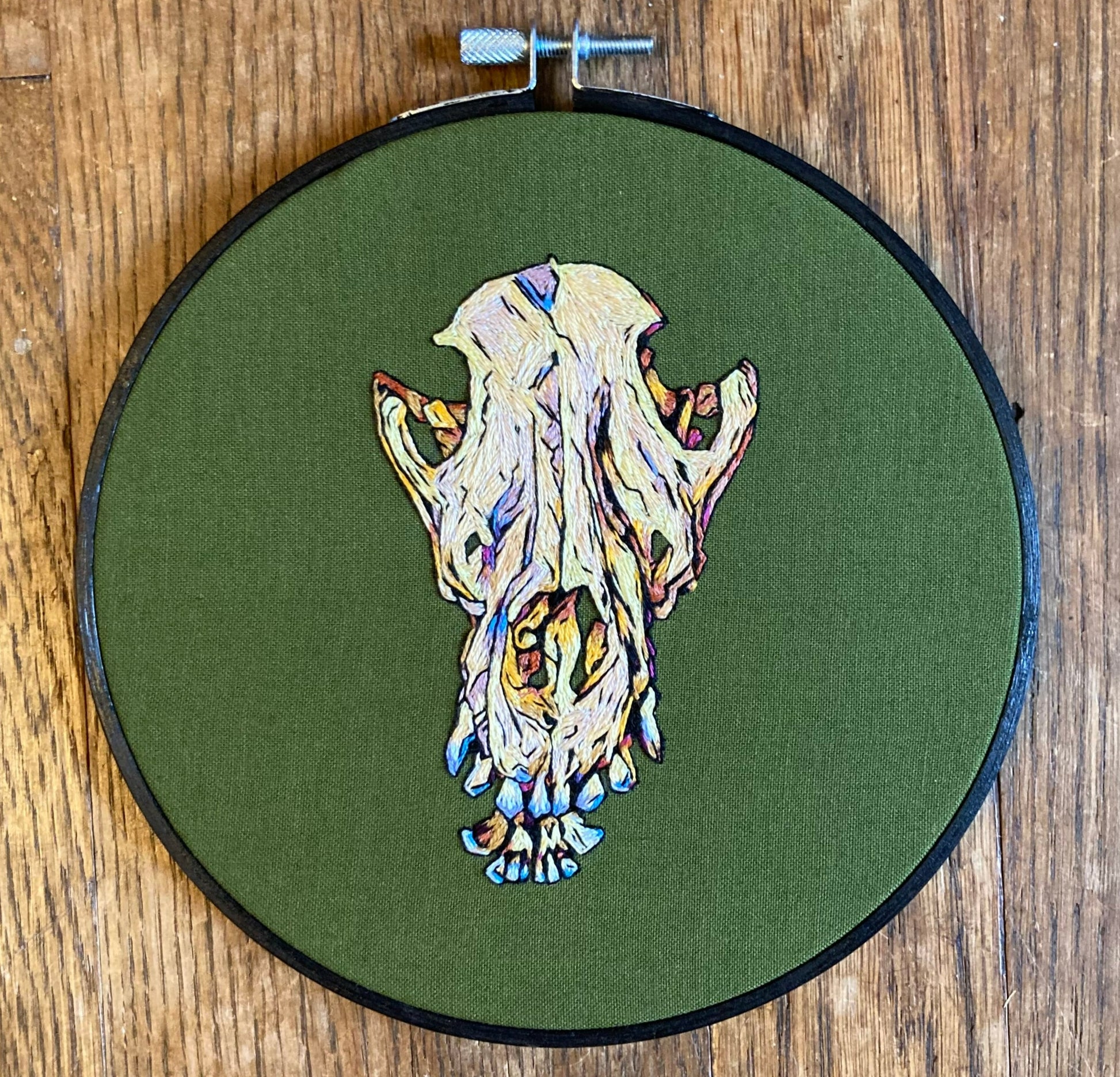 Colorful embroidery of a dog skull with prominent teeth on olive green fabric in a round embroidery hoop.