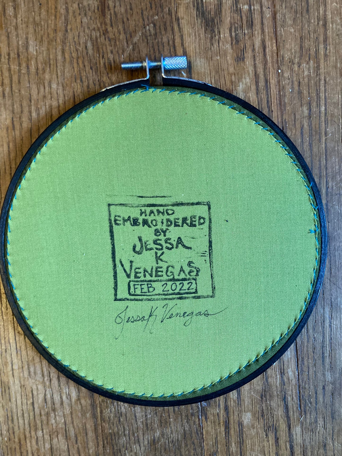 The back of an embroidery hoop with light green fabric stitched taut like a drum. A stamp reads "HAND EMBROIDERED BY JESSA K VENEGAS FEB 2022"